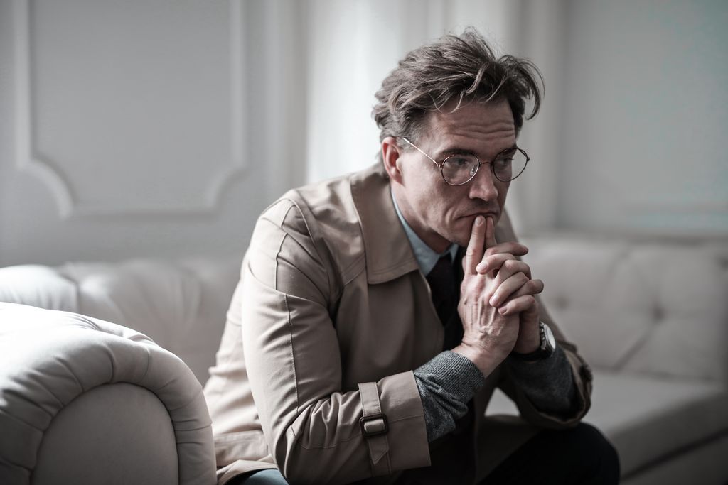A thoughtful man wearing glasses sits on a sofa in a classic interior, lost in deep contemplation.