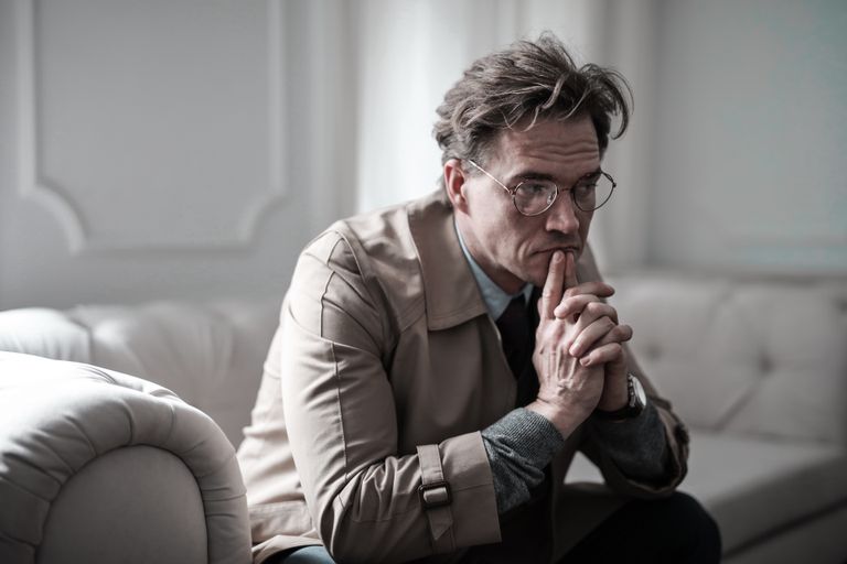 A thoughtful man wearing glasses sits on a sofa in a classic interior, lost in deep contemplation.